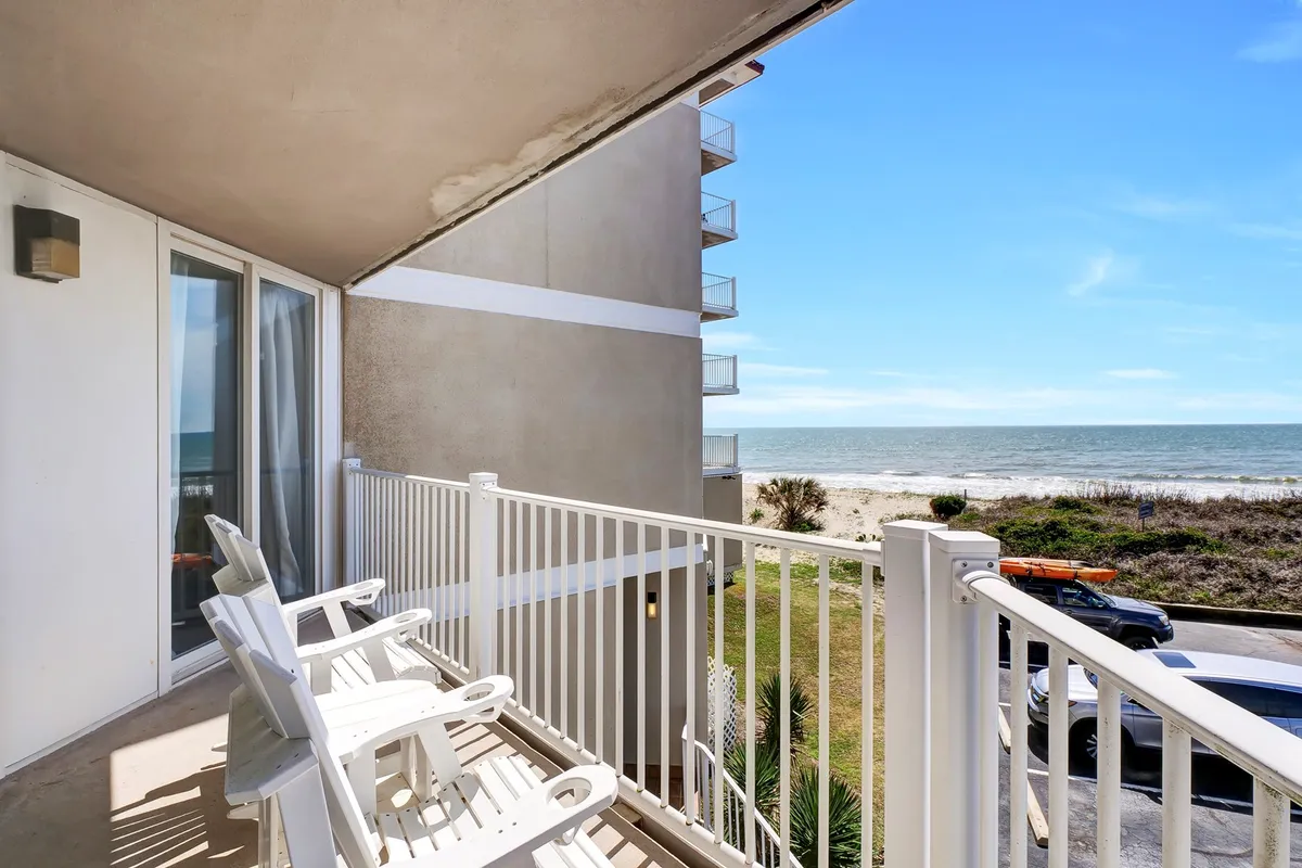 A private balcony with comfortable chairs overlooks the expansive ocean and stretches of sandy North Topsail Beach beneath a clear blue sky. The view invites guests to relax with a morning coffee or evening cocktail, with sunlight glinting off the gentle waves below.