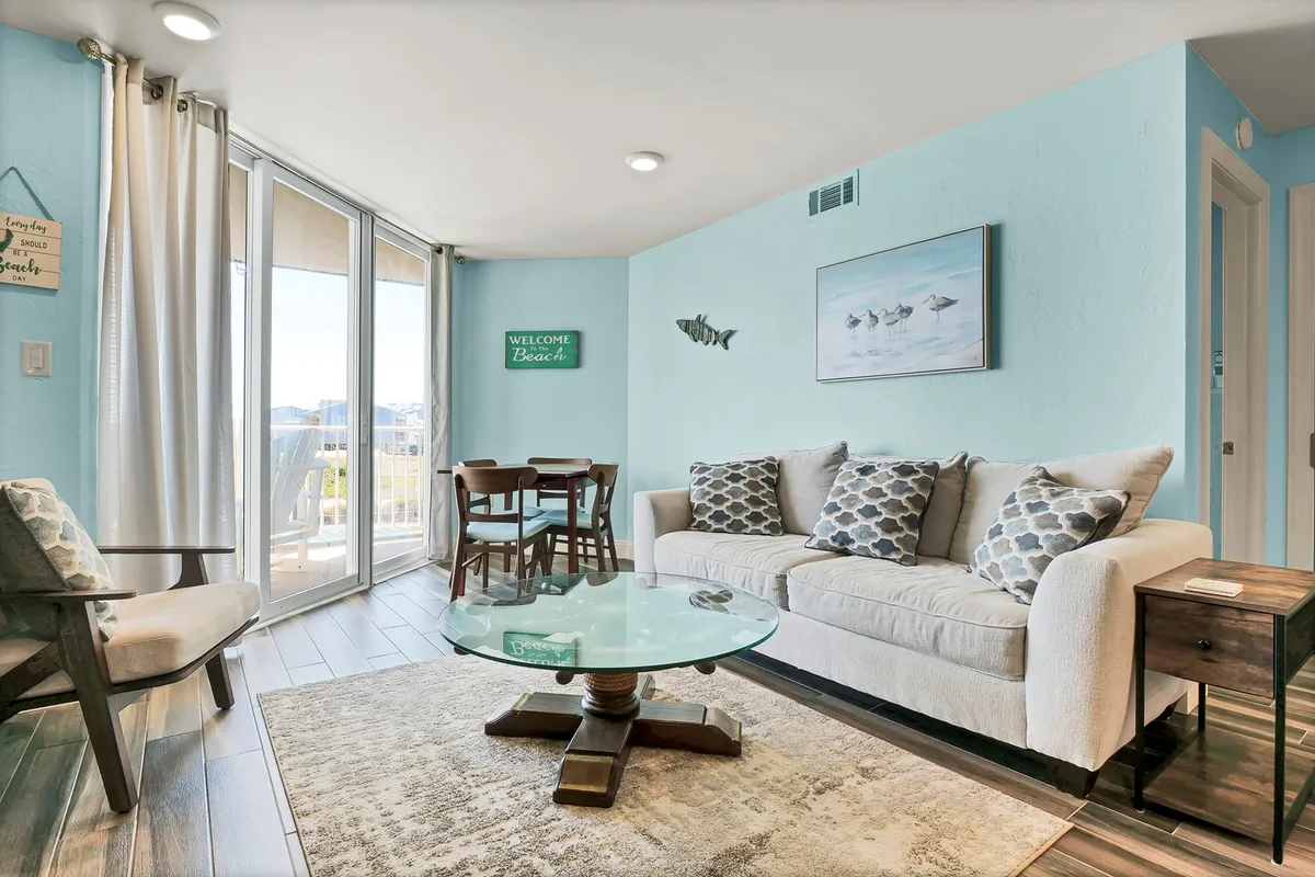 A bright living room and dining space are visible, featuring a plush sofa, coffee table, and dining table, all oriented towards a large window that offers an impressive ocean view. The ambiance is lighthearted and welcoming, with ample space for family or group activities.