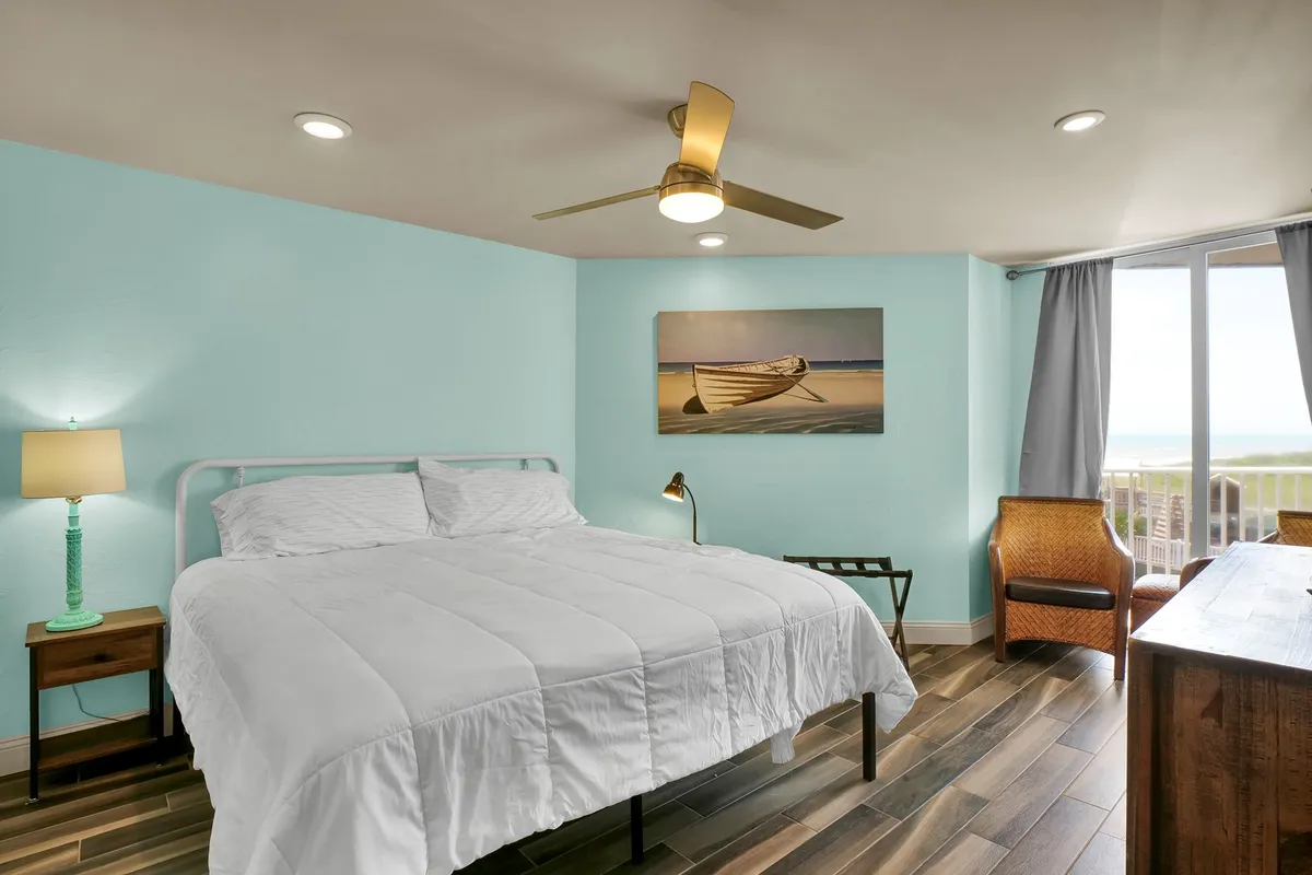 A tranquil master bedroom featuring a comfortable bed, a painting of a boat, a lamp, a wooden desk, and large windows revealing an expansive ocean view. The setting is serene and bright, ideal for restful mornings and evenings.