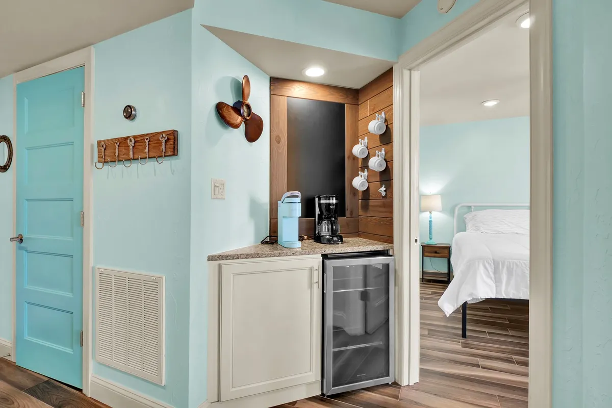 A cozy coffee station set beside a turquoise door, with wooden wall decor. In the background, a glimpse of an adjacent bedroom with fresh white bedding is visible, reinforcing the connection to the master suite. The scene feels intimate and perfect for starting the day with a warm drink.