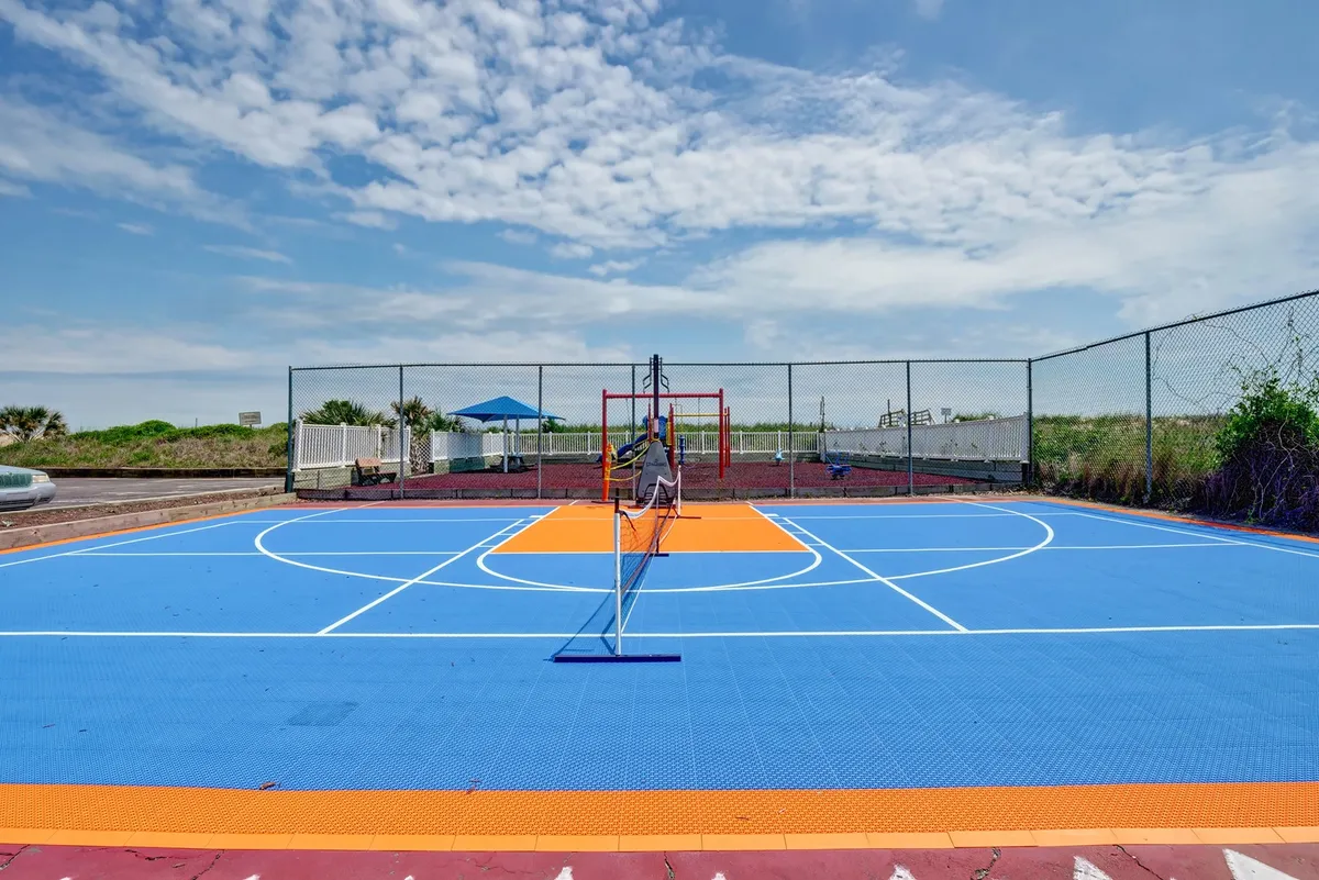 A colorful outdoor sports court with basketball hoops, a tennis net, and vibrant playground equipment in the background. Children are visible playing on the playground, and the scene radiates activity and energy.