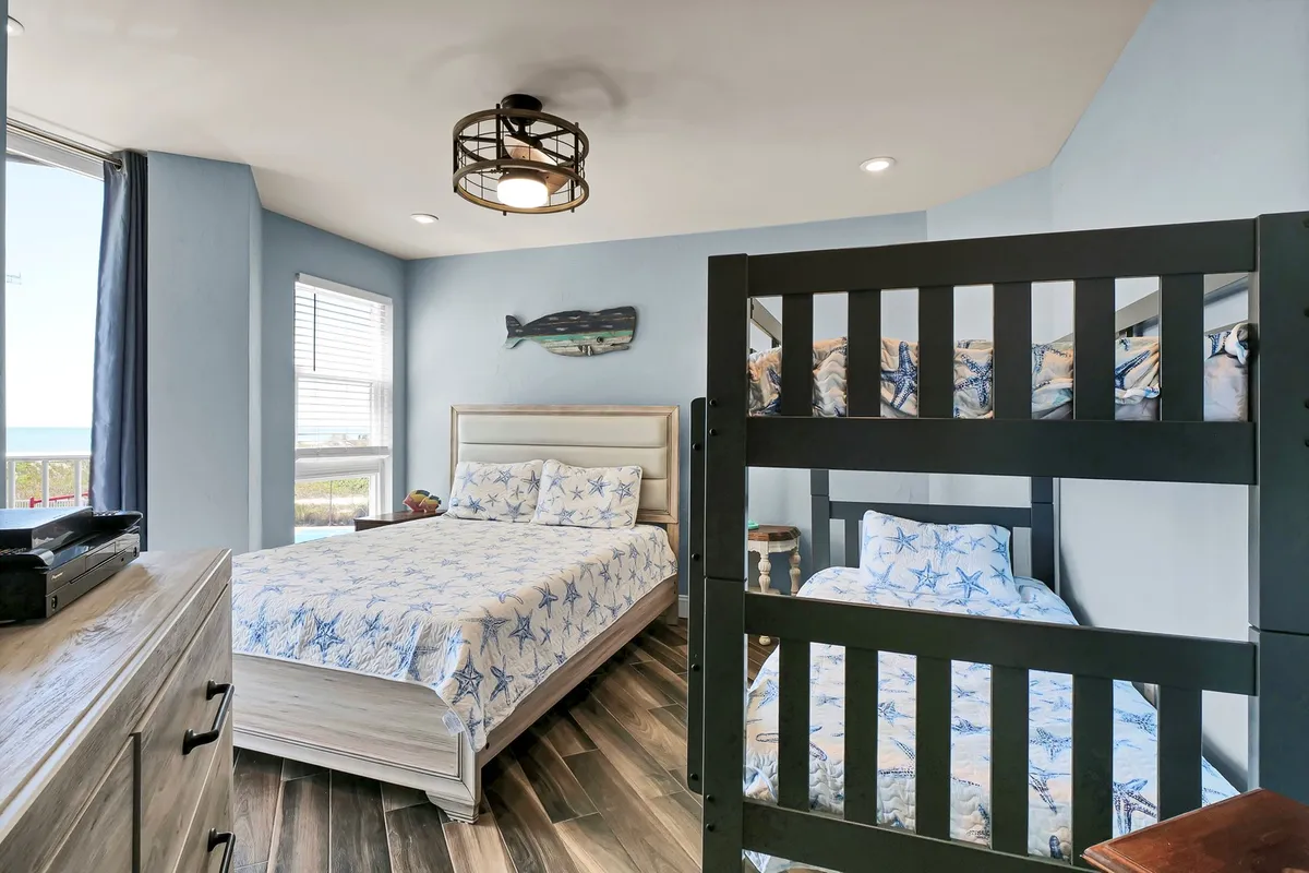 A cheerful and comfortable bedroom featuring a queen bed, twin-over-twin bunk, a decorative fish wall art, and a window offering ocean views. The room is bathed in natural light, creating a welcoming space for both relaxation and family bonding.