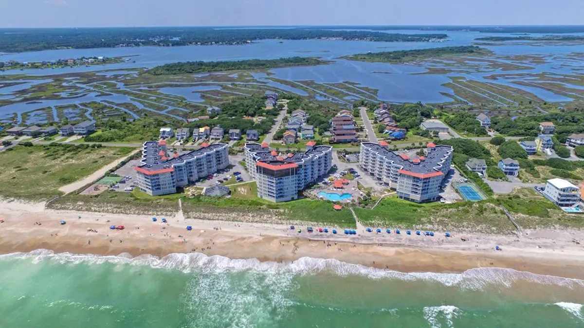An inviting view of beachfront buildings with a large swimming pool in the foreground, surrounded by lush landscaping and the sparkling sand and water of North Topsail Beach beyond.