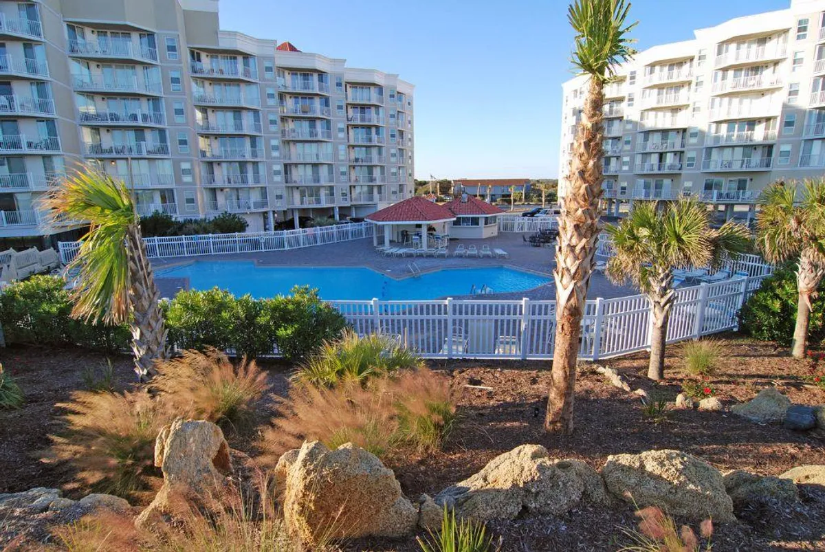This image displays a large, sparkling swimming pool surrounded by palm trees and resort buildings, with landscape rocks enhancing the tropical feel. The setting is bright and leisurely, suggesting a relaxing environment for swimming, sunbathing, and socializing.