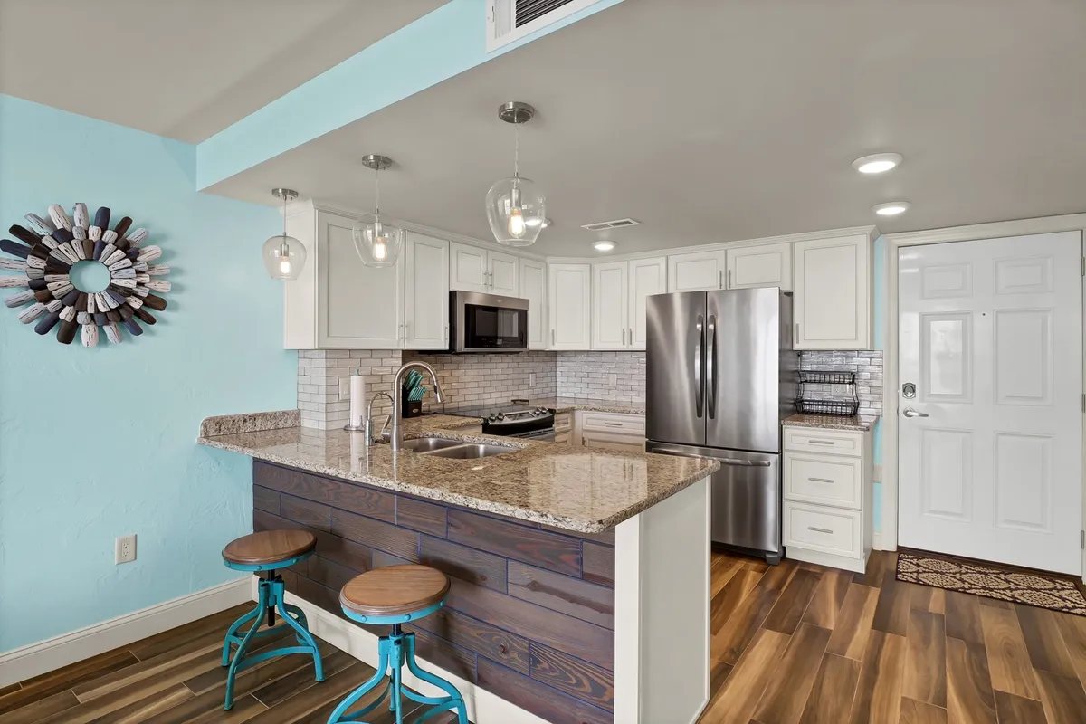 A bright and cheerful kitchen area with colorful bar stools lined up at a granite countertop, complemented by modern appliances and a lively, welcoming atmosphere. Decorative touches and abundant natural light make this space ideal for casual dining and socializing.