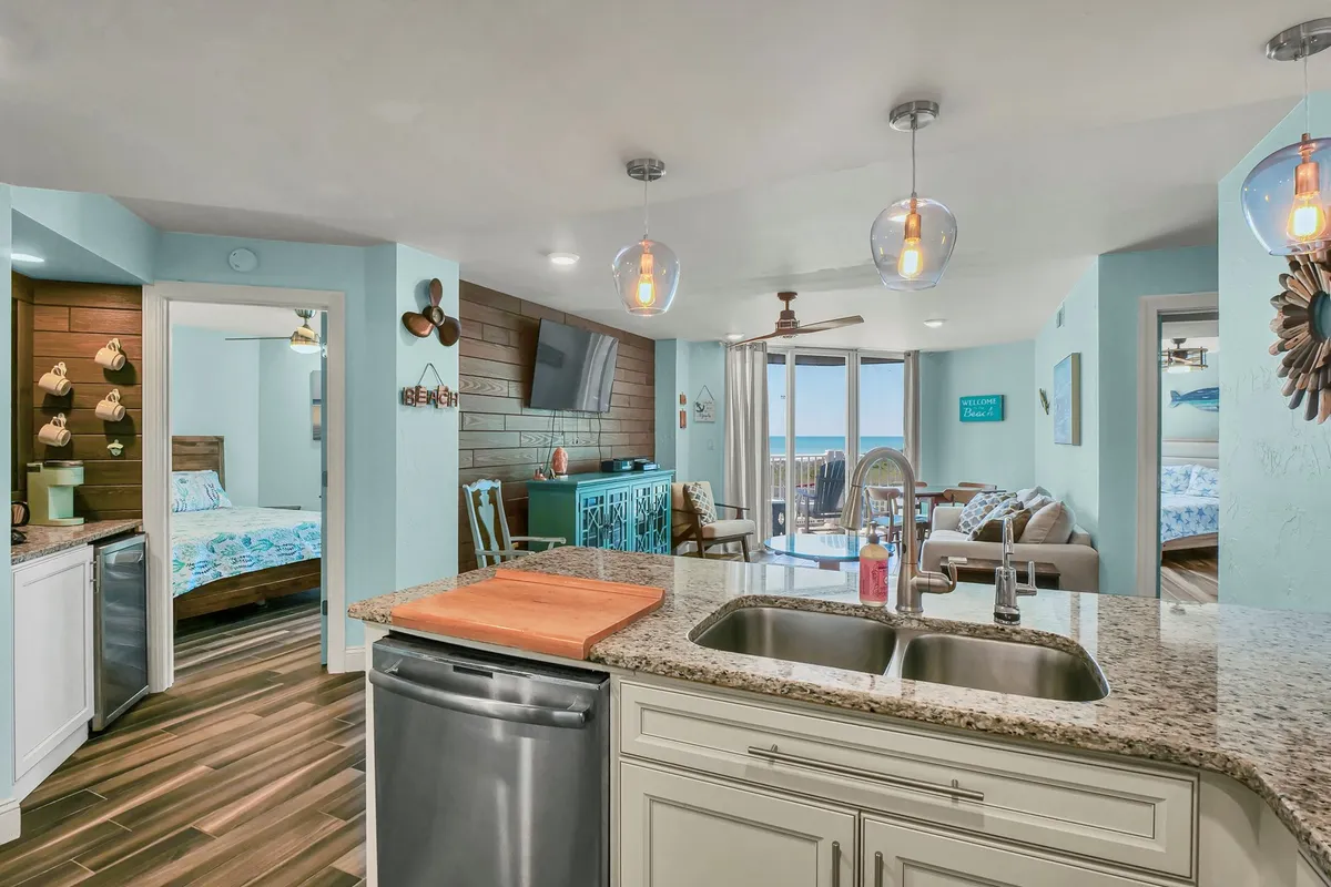 This image showcases an open-plan interior where the kitchen island, dining area, and living room all flow together. Large windows and a sliding glass door reveal an inviting ocean view and access to a private balcony, while natural light fills the space. The living room features comfortable seating and a clear view towards the ocean, creating a seamless indoor-outdoor connection.