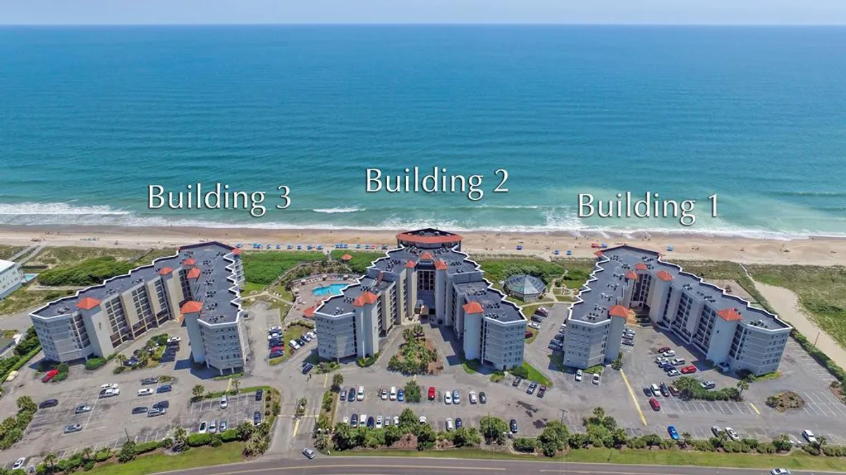 A sweeping view of three resort buildings situated beside a large outdoor swimming pool, with the sandy beachfront and ocean stretching in the background. Palm trees and lounge chairs create a welcoming, resort-style atmosphere.