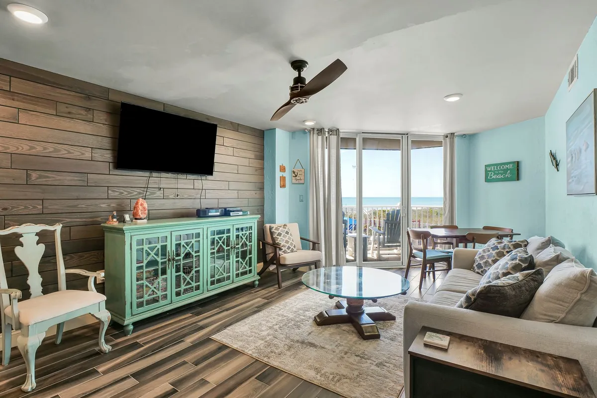 The image showcases a bright and inviting living area with a distinctive turquoise cabinet, plush seating arranged for relaxation, large windows that frame breathtaking ocean sunrises, and a warm wooden accent wall. The setup invites guests to unwind, enjoy scenic views, and gather for movie nights or special occasions.
