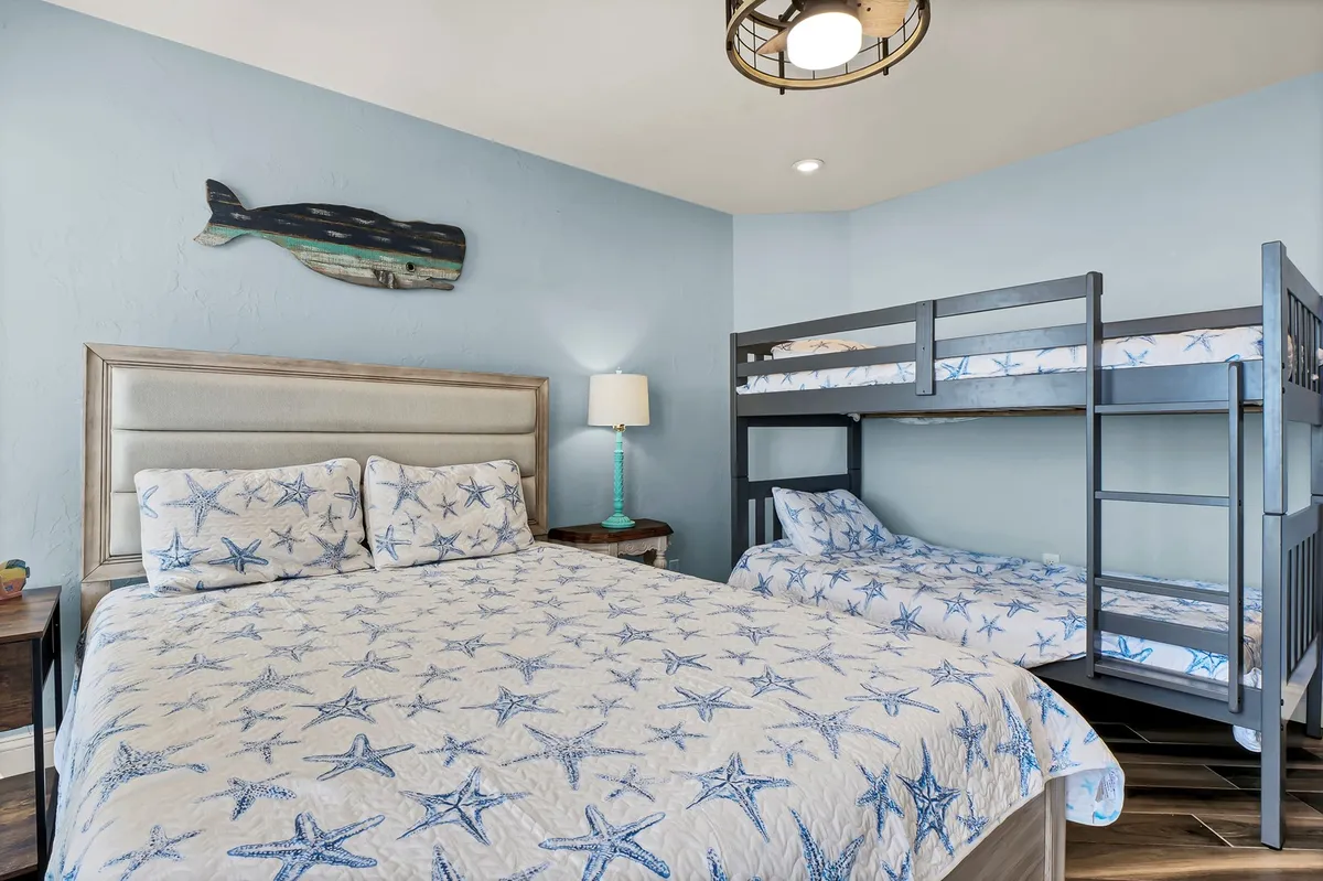 This image displays a cozy bedroom featuring a king-sized bed with starfish-patterned bedding, a twin-over-twin bunk bed, and decorative whale wall art, illuminated by a lamp with a turquoise base. The arrangement radiates warmth and family-friendly comfort.